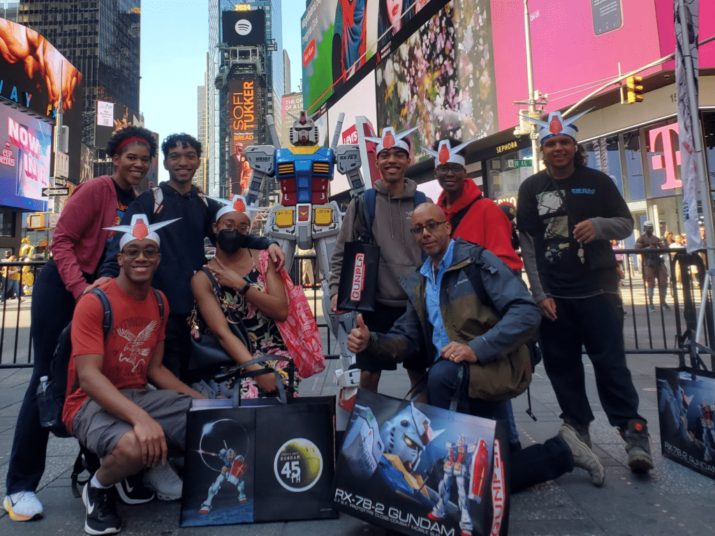 A group of people posing in front of a life size Gundam robot while they wear Gundam hats as part of a pop up shop in New York City organized by Pro Motion.